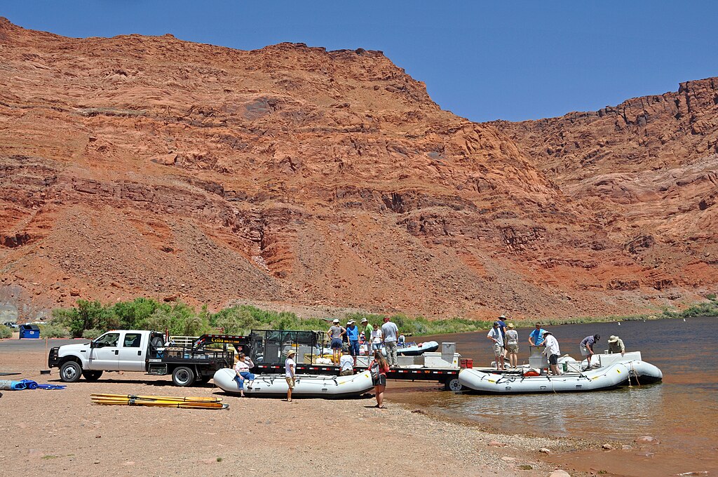 People prepare inflatable rafts by a river under a clear blue sky, with a white truck nearby. Rugged red cliffs rise in the background.