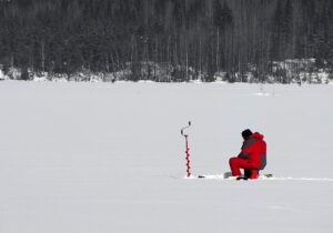 A person in a red snowsuit sits ice fishing on a frozen lake, using an ice auger. Snowy ground contrasts with dark forested background. Tranquil scene.