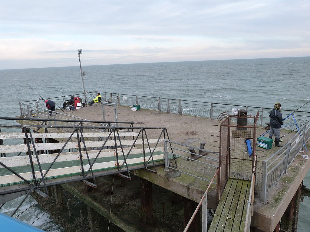 A small concrete pier extends into the ocean under a cloudy sky. Several people are fishing with rods, creating a peaceful, quiet atmosphere.