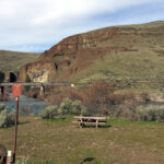 A peaceful landscape with a grassy field, a wooden picnic table, and a river flowing under a bridge. Rocky hills rise in the background under a blue sky.