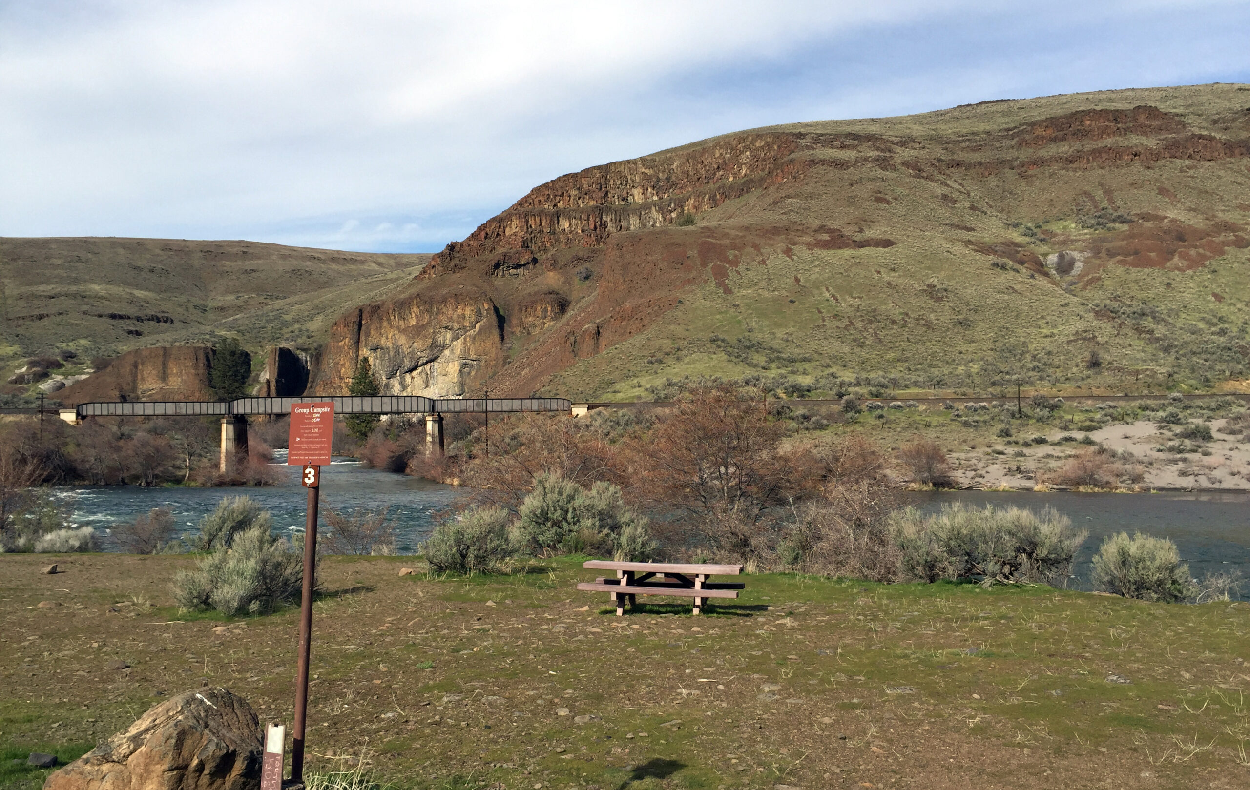 A peaceful landscape with a grassy field, a wooden picnic table, and a river flowing under a bridge. Rocky hills rise in the background under a blue sky.