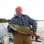 An elderly person in a plaid jacket and orange hat proudly holds a large fish on a boat. The background shows calm water and a wooded shoreline.