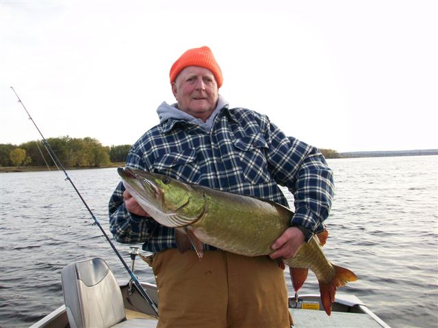 An elderly person in a plaid jacket and orange hat proudly holds a large fish on a boat. The background shows calm water and a wooded shoreline.