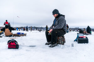 A man in winter clothing sits on a stool ice fishing, holding a small rod over a hole. Nearby, others are scattered across the snowy landscape, creating a quiet, communal atmosphere.