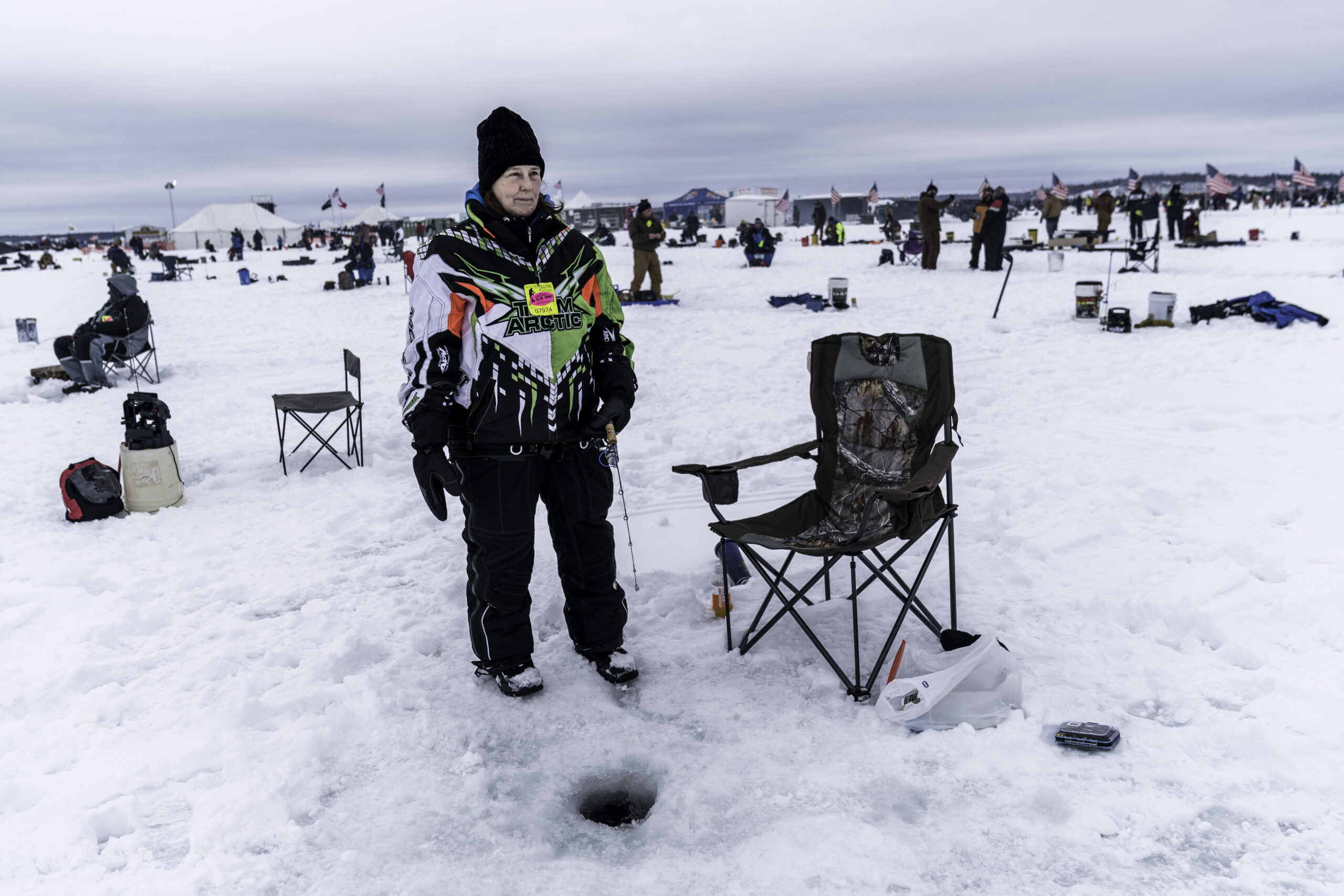 Person ice fishing, standing on a snowy lake. They wear a colorful jacket and black hat, next to a camo chair, with an ice hole nearby.