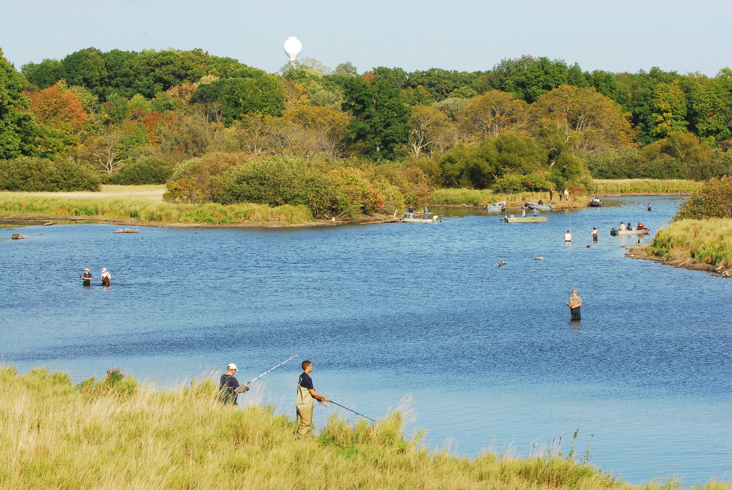 A serene river scene with people fishing under a clear blue sky. Trees in early autumn colors surround the water. The mood is peaceful and calm.