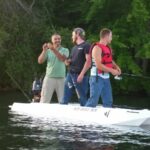 Three men are standing on a small white boat, fishing and laughing. One man holds a fishing line, while the others watch. Green trees and calm water are in the background.