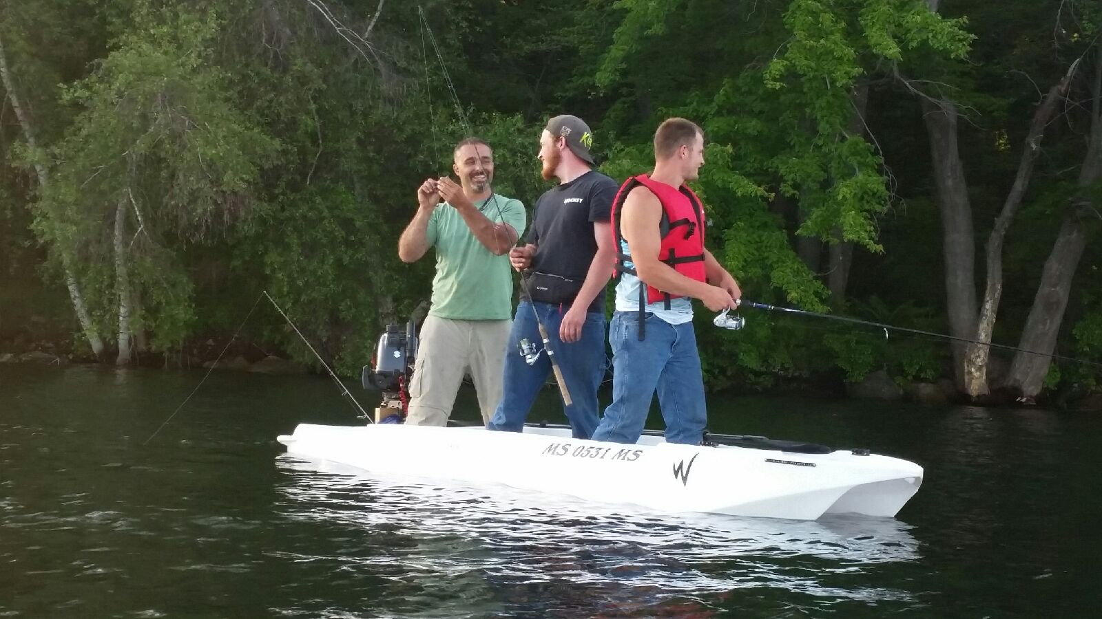 Three men are standing on a small white boat, fishing and laughing. One man holds a fishing line, while the others watch. Green trees and calm water are in the background.