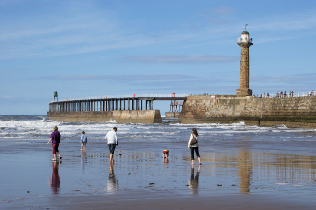 People walk along a reflective wet beach near a pier and tall lighthouse under a clear blue sky. The scene is calm and bright, with gentle waves.