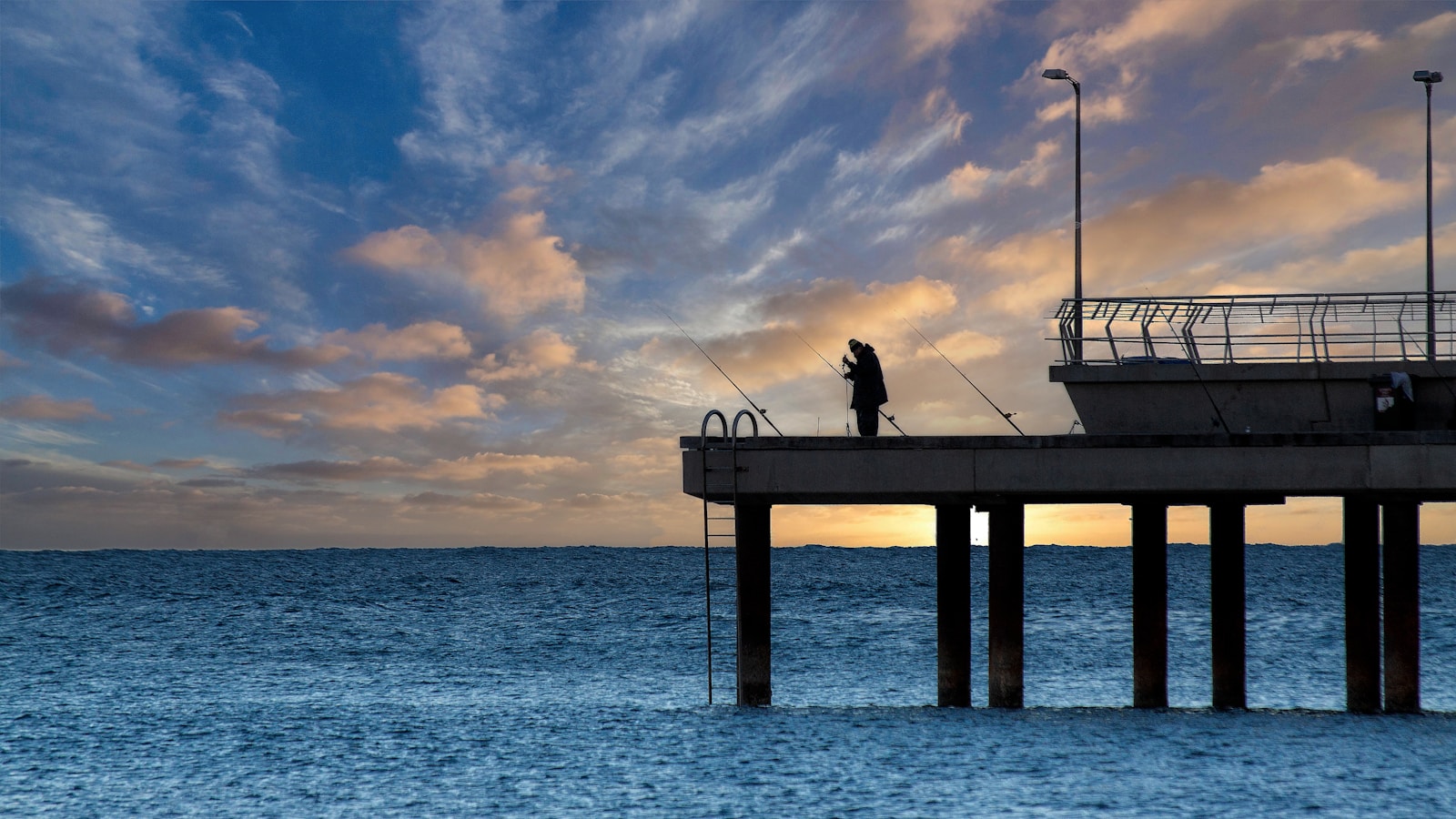 A man standing on a pier fishing at sunset.
