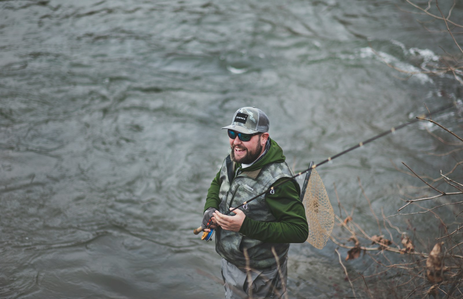 A man smiling, holding a black fishing rod near a body of water.