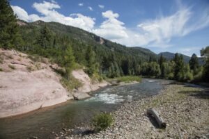 A serene river flows beside rocky shorelines and lush green trees, nestled in a forested mountain landscape under a blue sky with scattered clouds.