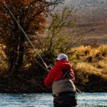 A person in waders and a red jacket fly fishing in a river during autumn, with trees and dry grass on the banks.